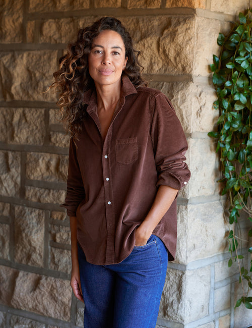 A woman with curly brown hair wearing a brown corduroy button-down shirt and blue jeans, standing against a stone wall with green ivy on the right., front view