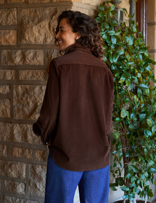 A woman wearing a dark brown long-sleeve button-up shirt and blue jeans, standing in front of a stone wall next to a green potted plant., alternate view