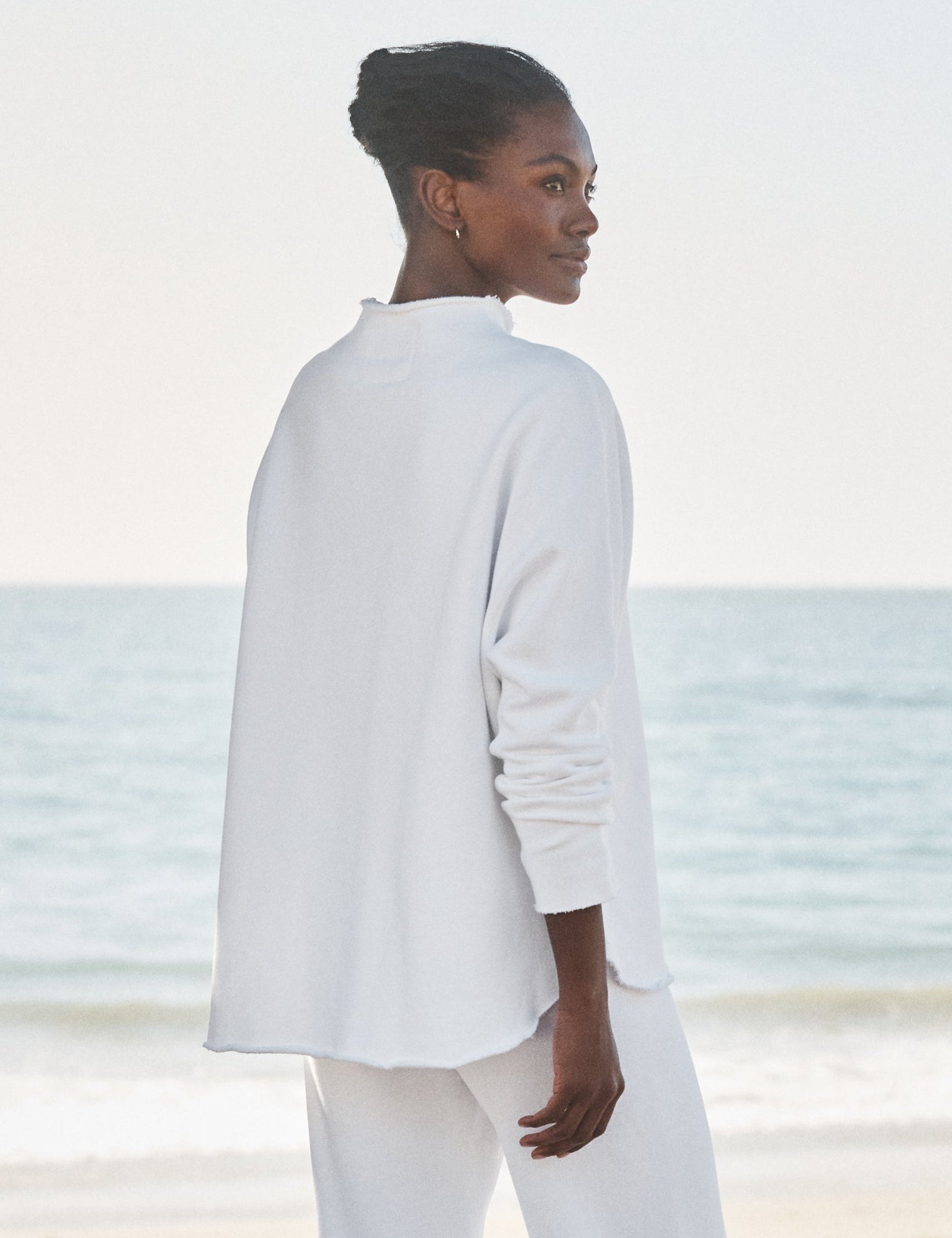 Person wearing a white outfit standing on a beach with ocean in the background, alternate view