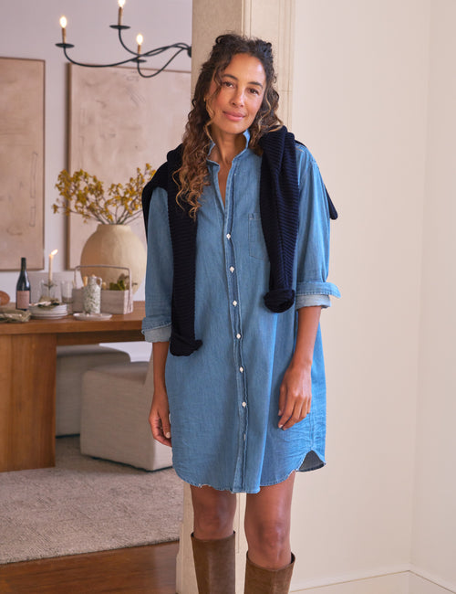 Woman wearing a blue dress with a navy sweater over her shoulders in a room with a wooden table and decor., front view