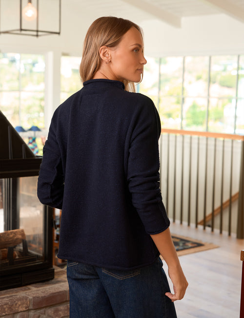 A woman wearing a dark navy blue pullover sweater and dark blue jeans, shown from the back in a bright, modern home interior., alternate view