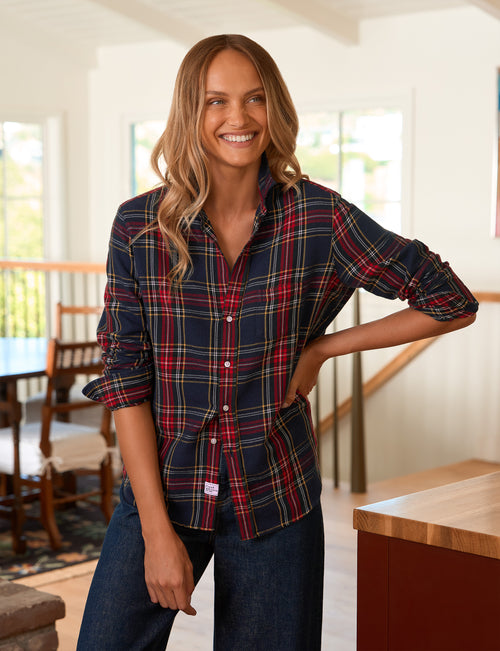 A woman modeling a long-sleeve button-up plaid shirt in a home interior setting. The shirt features a navy blue base with red, green, and yellow plaid stripes., front view