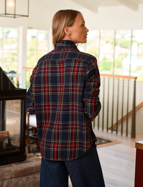 A woman wearing a navy blue plaid flannel shirt with red, yellow, and white stripes, shown from the back in a bright indoor home setting., alternate view