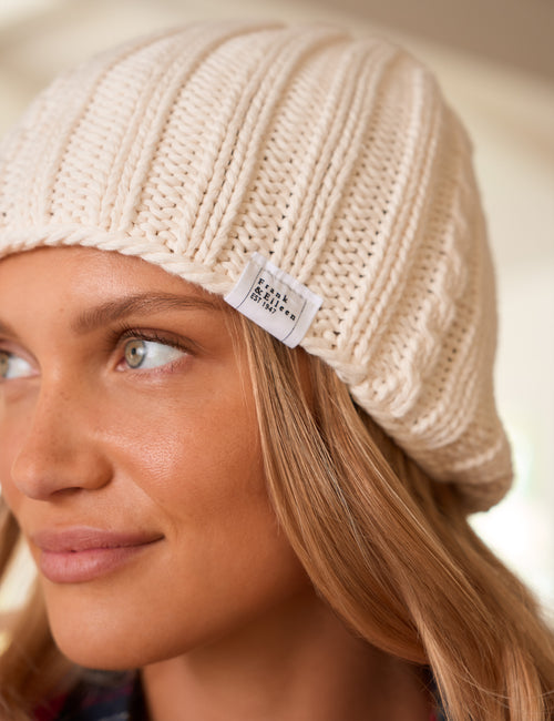 Close-up of a woman wearing a cream-colored ribbed knit beanie with a small white fabric label that reads 'Frank & Eileen'., alternate view