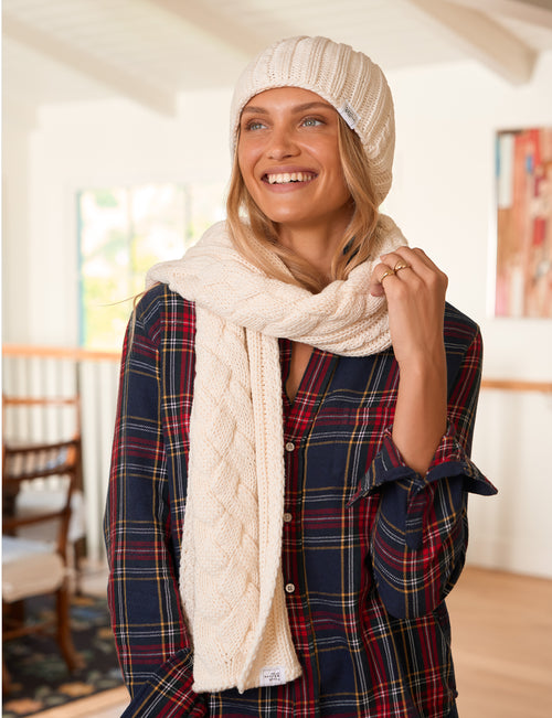 A smiling woman wearing a cream cable-knit beanie, matching knit scarf, and a navy red plaid flannel shirt in a bright indoor setting., front view