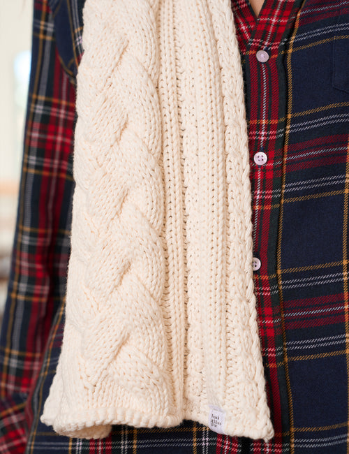 A close-up of a cream cable-knit scarf draped over a navy and red plaid button-up shirt., alternate view