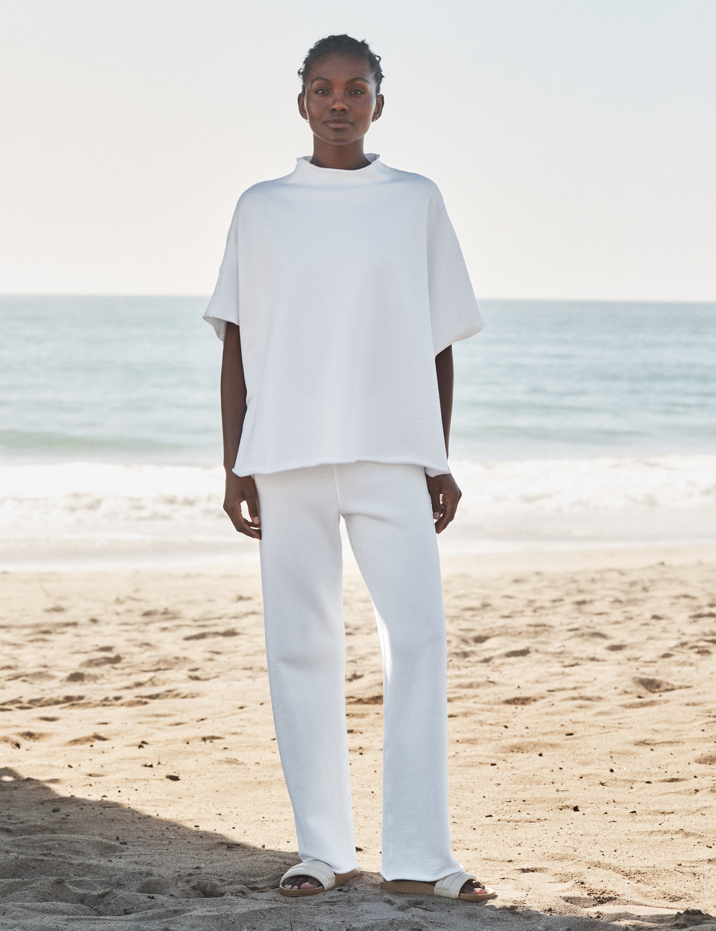 Person wearing a white outfit standing on a beach with ocean in the background, front view