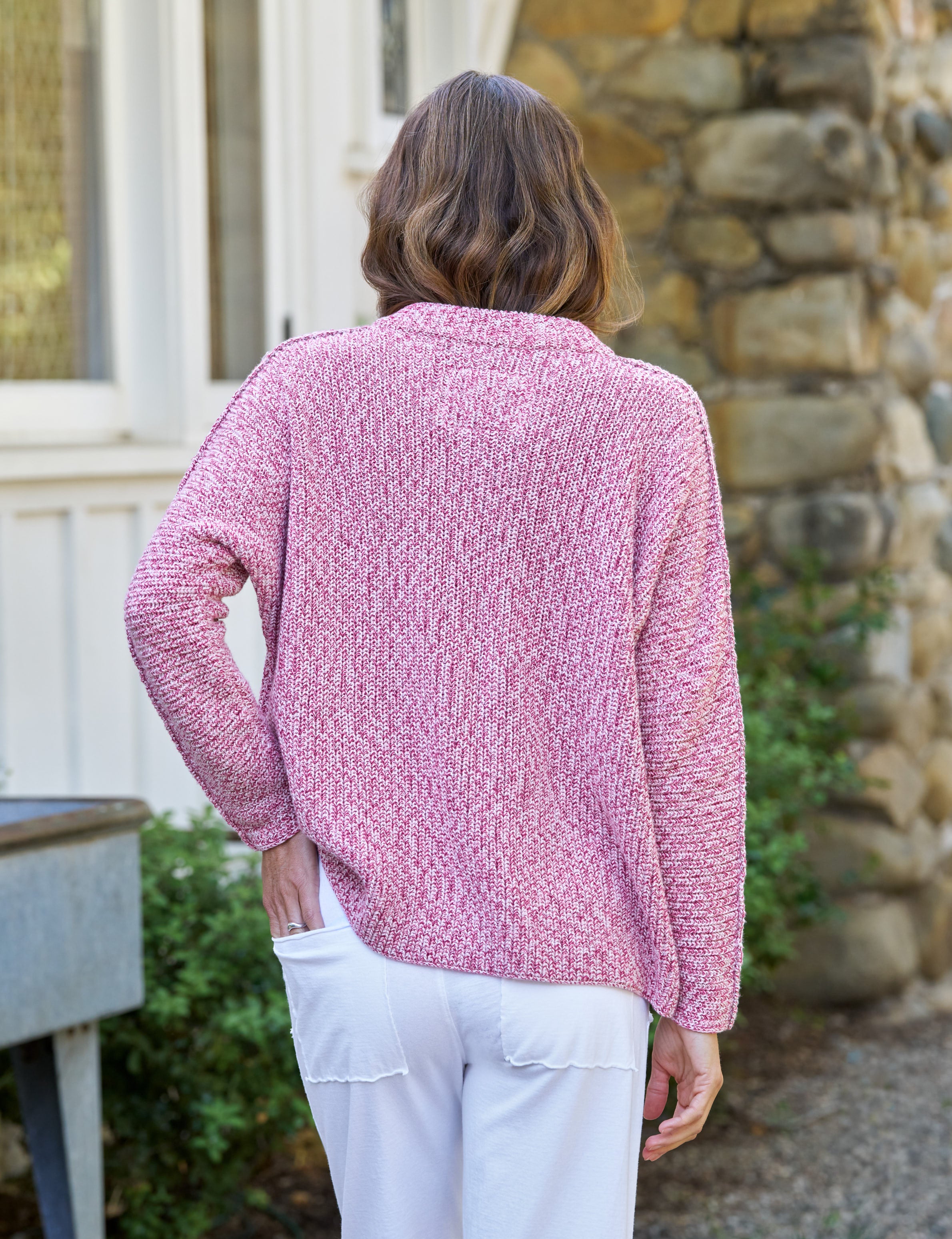 Woman wearing a pink textured sweater and white pants standing outdoors., alternate view