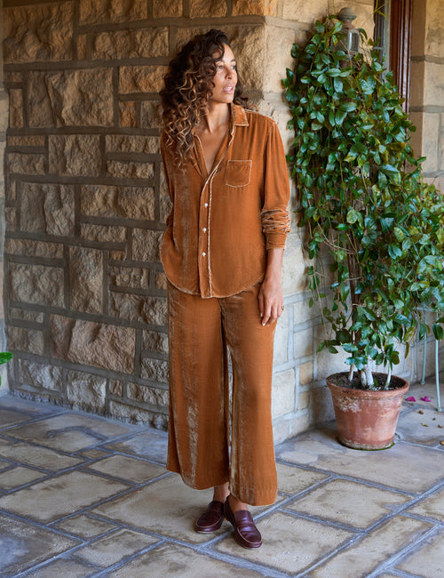 A woman wearing a gold two-piece set consisting of a button-down shirt and matching wide-leg pants, standing on a stone patio next to a potted green plant., view 4
