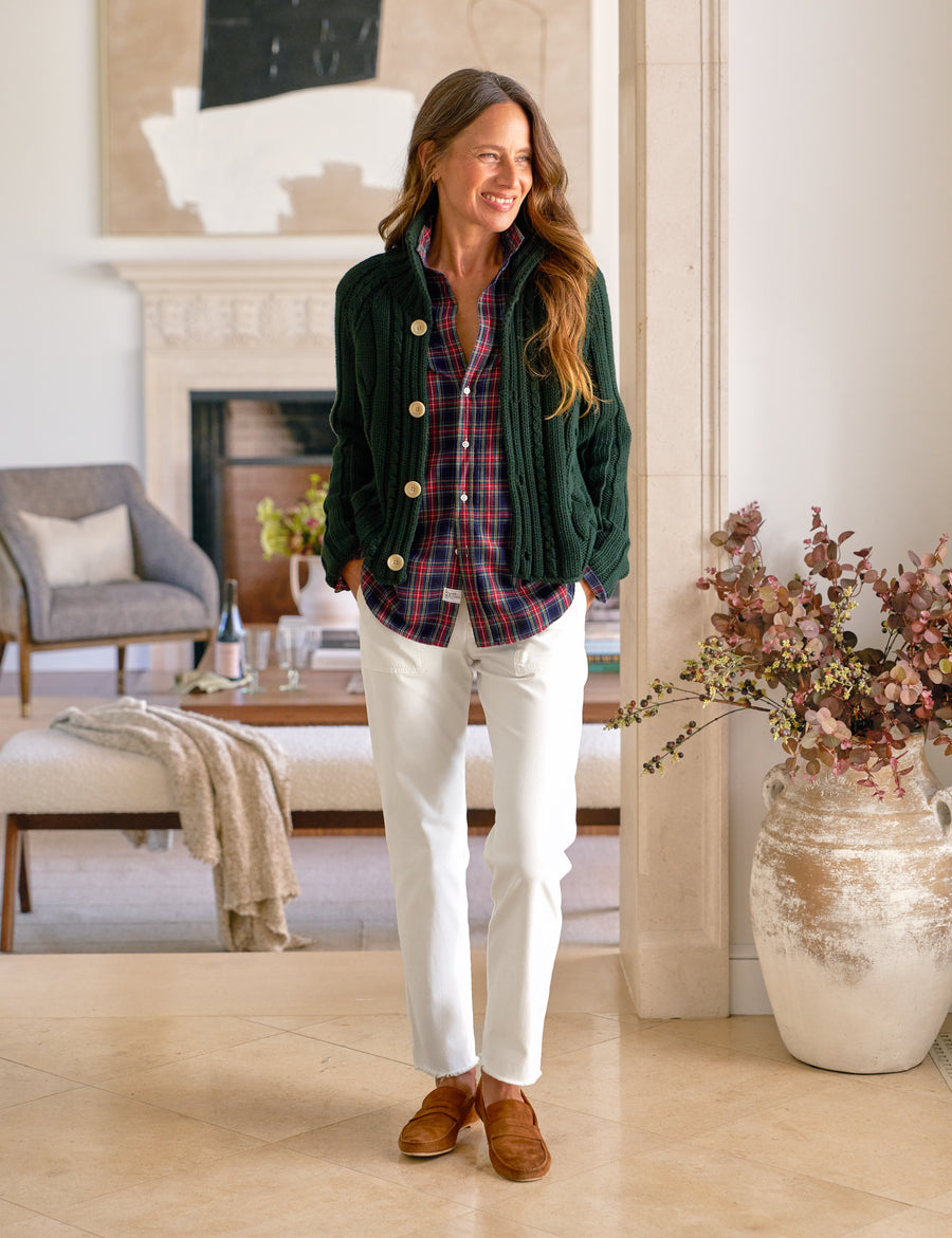 Woman standing in a living room wearing a green jacket, plaid shirt, and white pants.