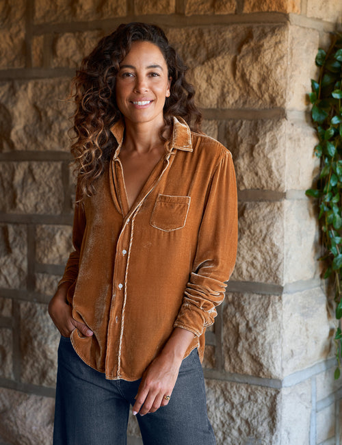 A woman with long curly brown hair stands outdoors against a stone wall, wearing a gold velvet button-up shirt with dark blue jeans., front view
