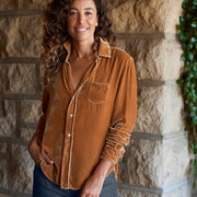 A woman with long curly brown hair stands outdoors against a stone wall, wearing a gold velvet button-up shirt with dark blue jeans.