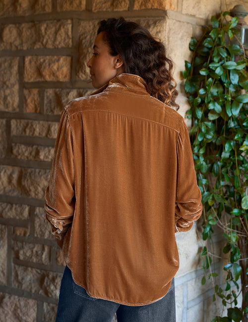 A woman wearing a gold velvet long-sleeve button-up shirt and blue jeans, standing in front of a stone wall with green ivy., alternate view