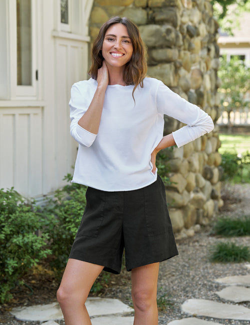 Woman wearing a white long-sleeve top and black shorts standing outdoors with a stone wall and greenery in the background., view 4