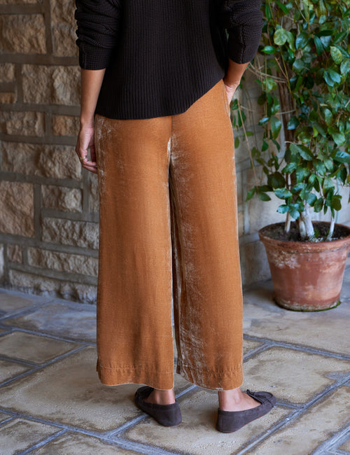 A person wearing wide-leg cropped pants in gold, styled with a brown ribbed long-sleeve sweater and dark slip-on shoes, standing on a stone patio next to a potted plant., alternate view