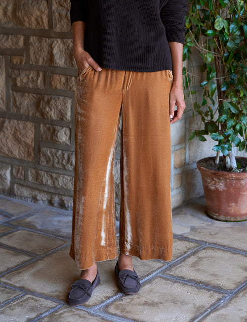 A person wearing wide-leg gold pants, a black ribbed long-sleeve sweater, and dark loafers, standing on a stone tile floor in front of a stone wall with a potted plant., front view