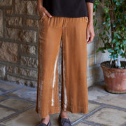 A person wearing wide-leg gold pants, a black ribbed long-sleeve sweater, and dark loafers, standing on a stone tile floor in front of a stone wall with a potted plant.