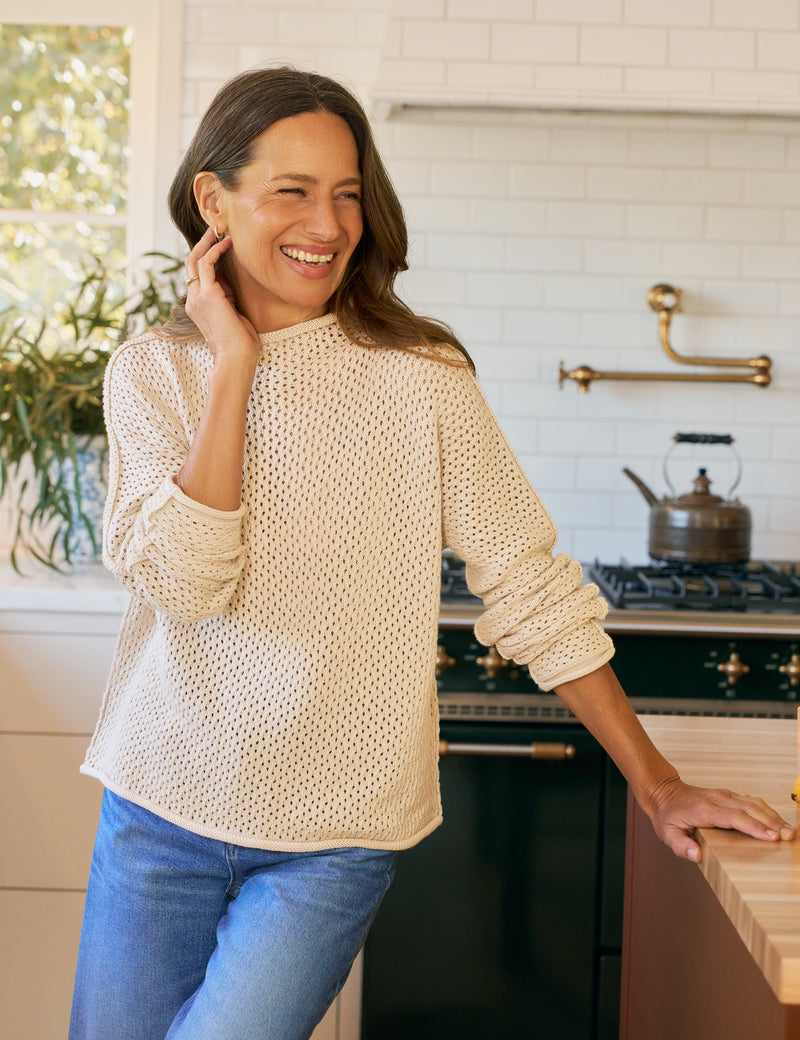 Woman standing in a kitchen wearing a beige sweater and blue jeans.