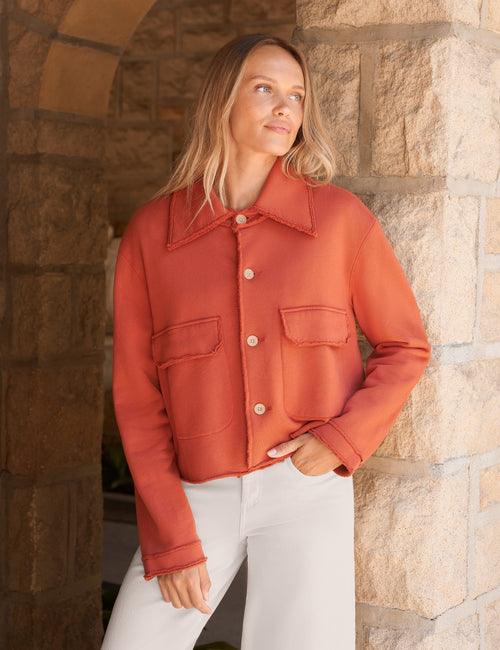 A woman wearing a cropped, rust-orange button-up jacket with a pointed collar and flap chest pockets, paired with white pants, standing in front of a rustic stone archway., front view
