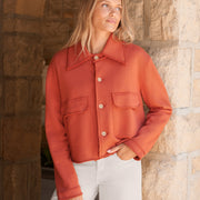 A woman wearing a cropped, rust-orange button-up jacket with a pointed collar and flap chest pockets, paired with white pants, standing in front of a rustic stone archway.