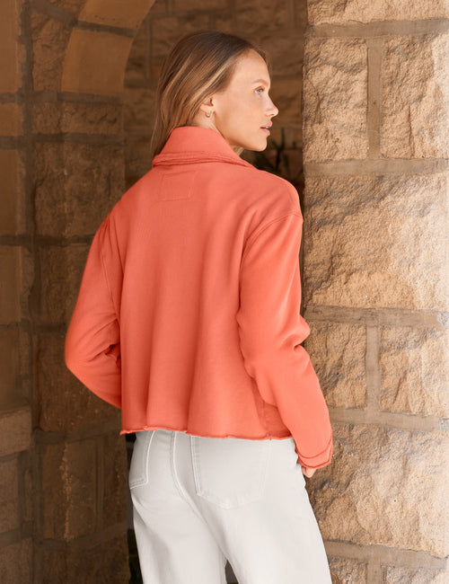 A woman wearing a orange cropped long-sleeve jacket and white pants, standing in profile in front of a rustic stone archway., alternate view