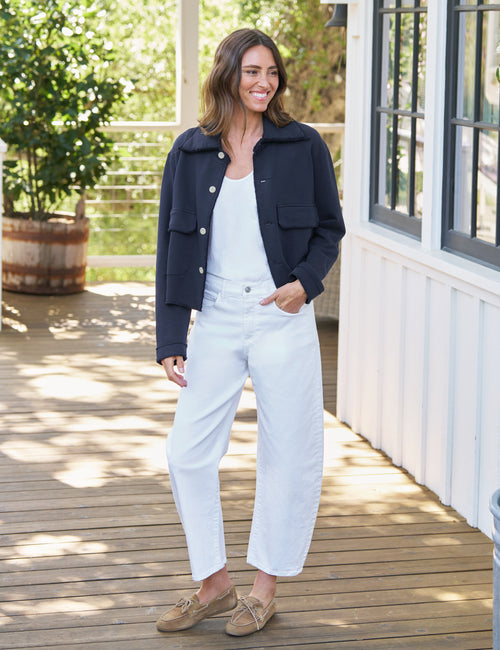 A woman stands on a wooden deck wearing a dark navy cropped jacket over a white tank top and white wide-leg pants, with tan loafers. The background shows a white house exterior, windows, and greenery., view 4