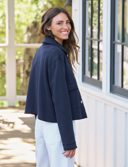 A woman wearing a navy blue cropped jacket and white pants, smiling and looking back over her shoulder in front of a white building with large windows., alternate view