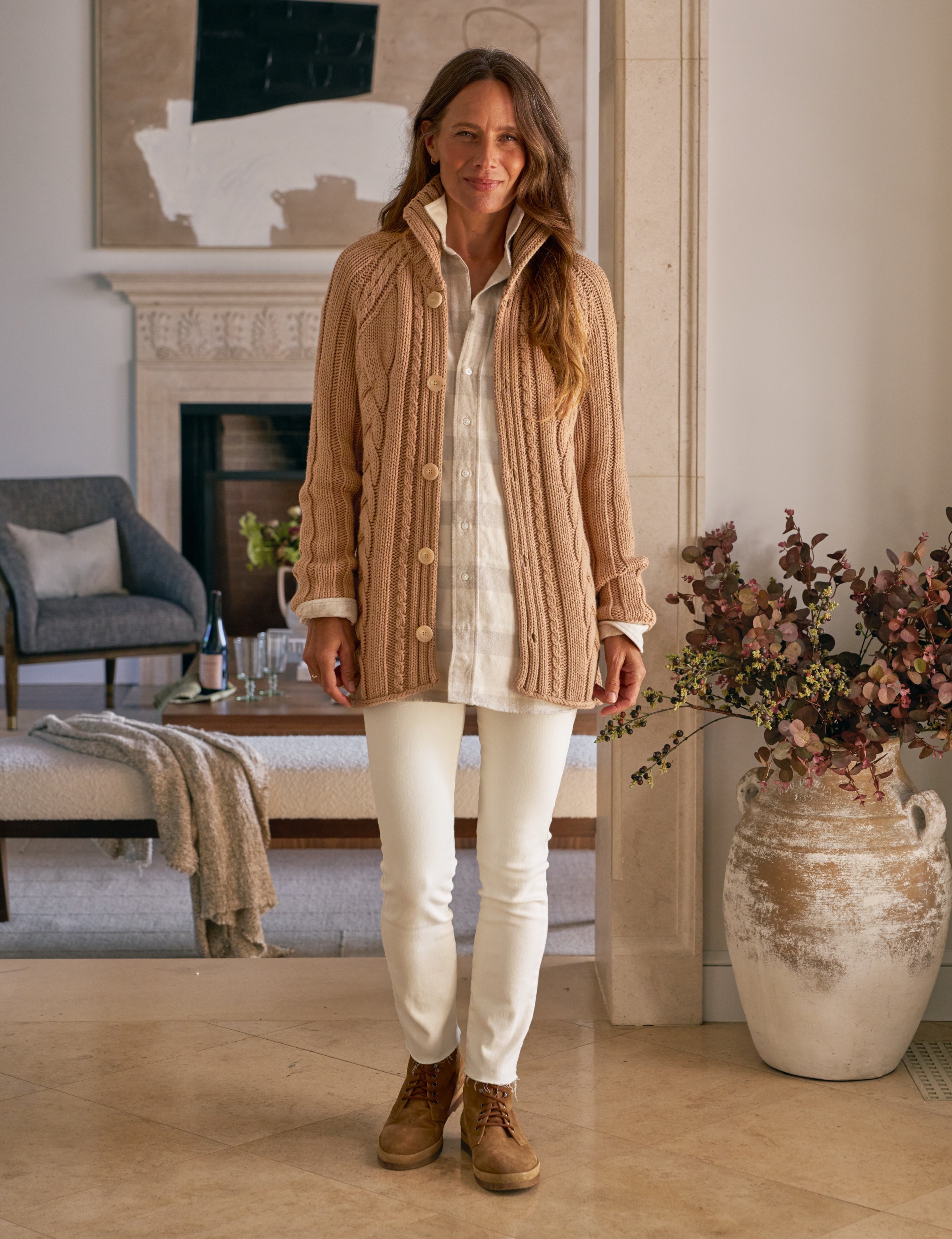 Woman standing in a living room wearing a beige cardigan, white pants, and brown boots.