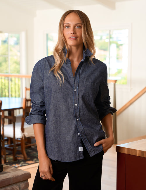 A woman wearing a dark blue  glitter chambray button-down shirt with rolled-up sleeves and black pants, standing in a bright, modern indoor setting with a staircase and dining area in the background., front view