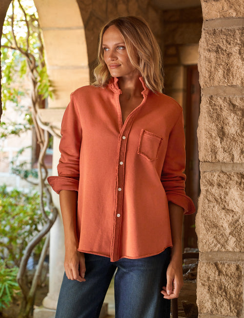 A woman wearing a rust-orange long-sleeve button-up shirt with a chest pocket, paired with dark blue jeans, standing in a rustic stone corridor., front view
