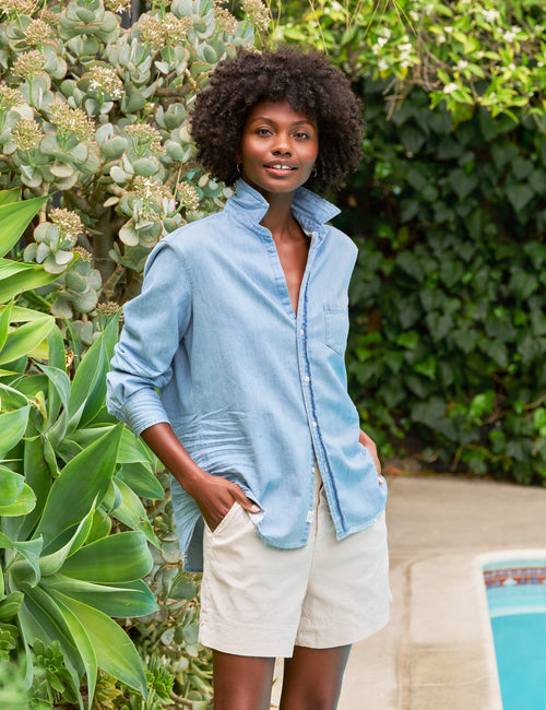 A woman standing outdoors wearing a classic blue button-up shirt and beige shorts, with a pool and greenery in the background., front view