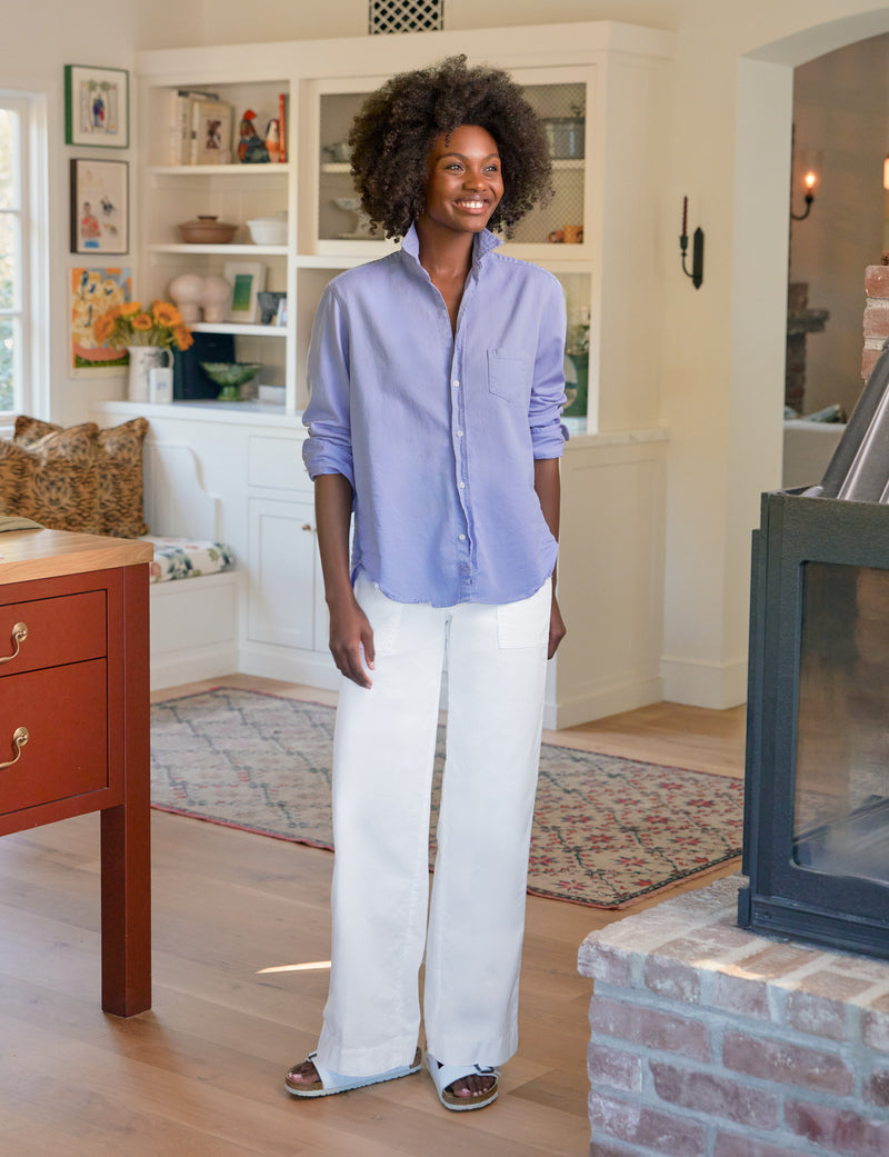 Woman standing in a living room wearing a light purple shirt and white pants.