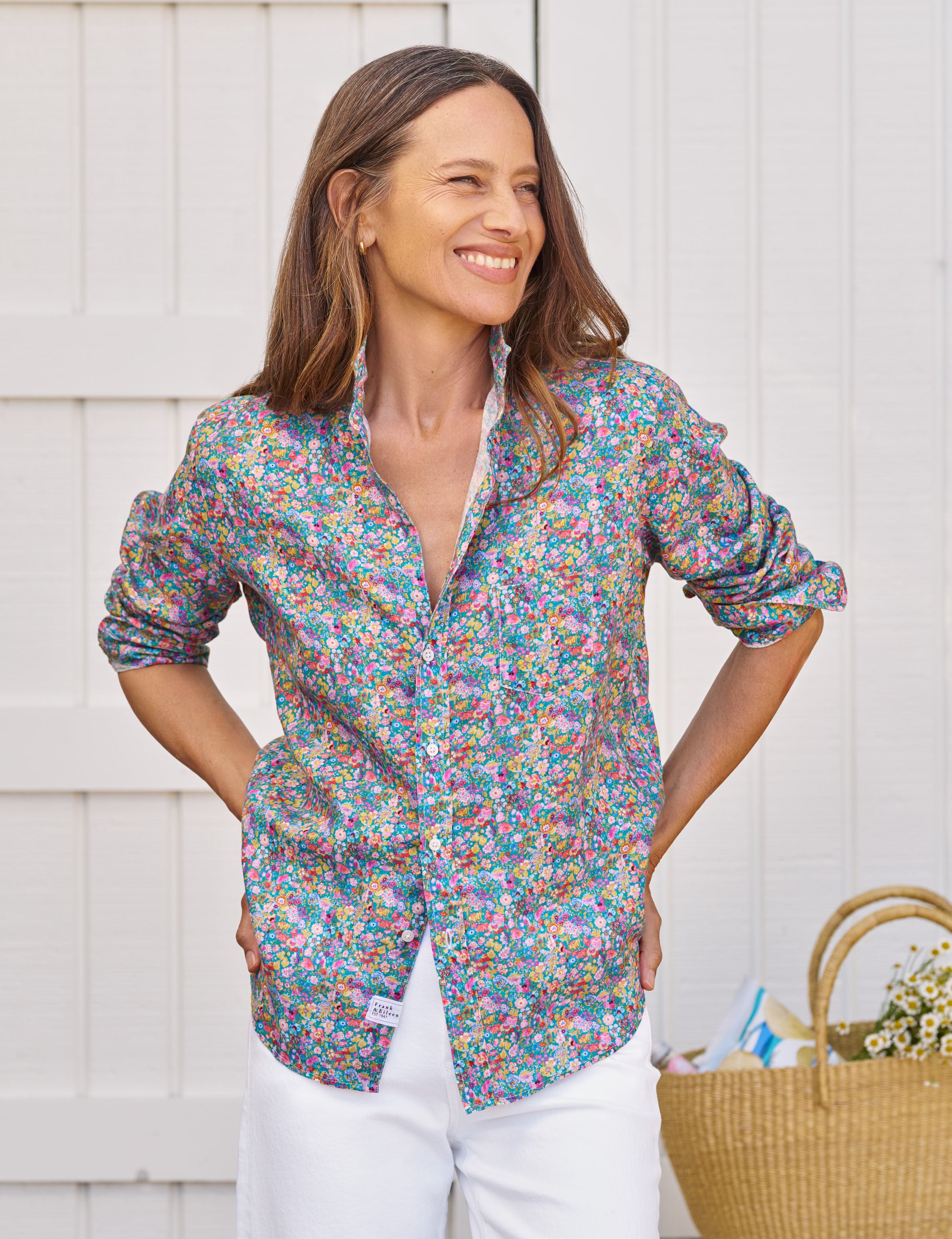 Woman wearing a colorful floral shirt with white pants standing against a light wooden wall., front view