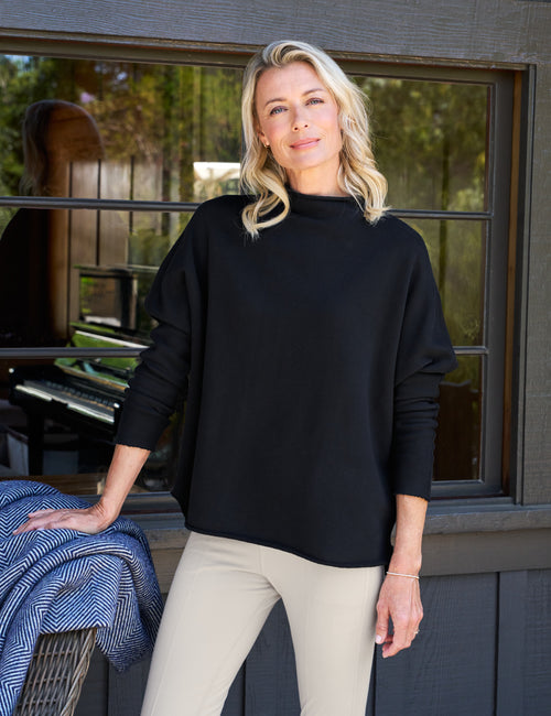 Woman wearing a black sweater and beige pants standing in front of a window with a view of trees., front view