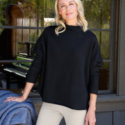 Woman wearing a black sweater and beige pants standing in front of a window with a view of trees.