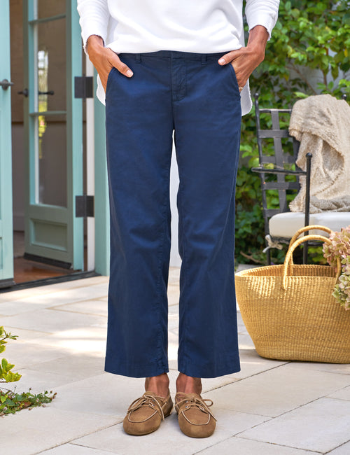 Person wearing navy pants and brown shoes standing outdoors with a wicker basket and chair in the background., front view