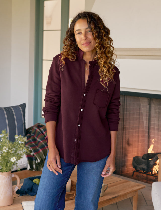 Woman wearing a burgundy shirt and blue jeans standing in a cozy living room with a fireplace.