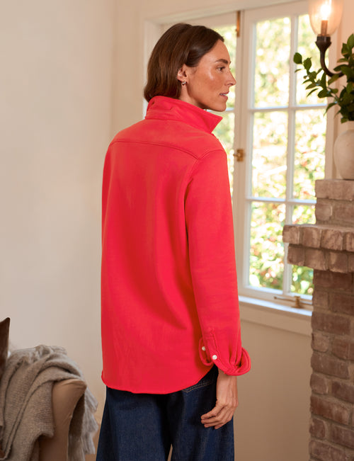 A woman wearing a bright red long-sleeve shirt and dark blue jeans, standing in profile in a cozy indoor setting with a window and brick fireplace visible., alternate view