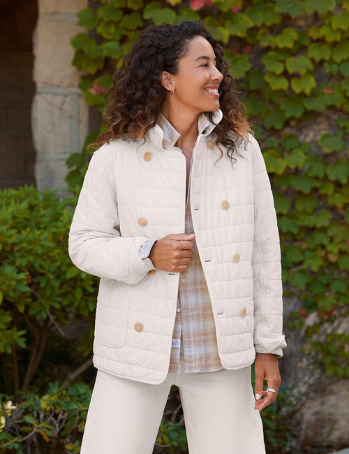 A woman wearing a cream quilted jacket with tan buttons, styled over a plaid shirt and cream pants, standing outdoors in front of a stone wall and green ivy., front view