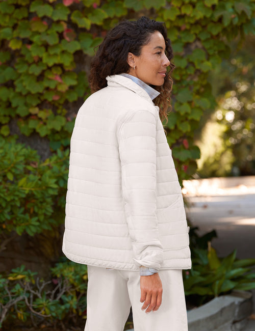 A woman wearing a white quilted jacket and matching white pants, standing outdoors in front of a leafy green wall., alternate view