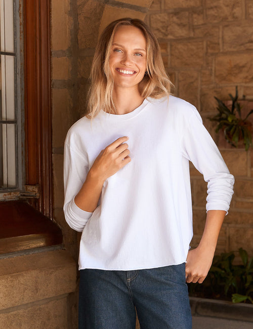 A woman stands on a stone porch wearing a white long-sleeve crewneck top, dark blue wide-leg trousers, and dark brown leather shoes., front view