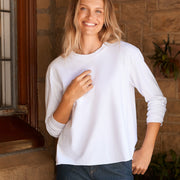 A woman stands on a stone porch wearing a white long-sleeve crewneck top, dark blue wide-leg trousers, and dark brown leather shoes.