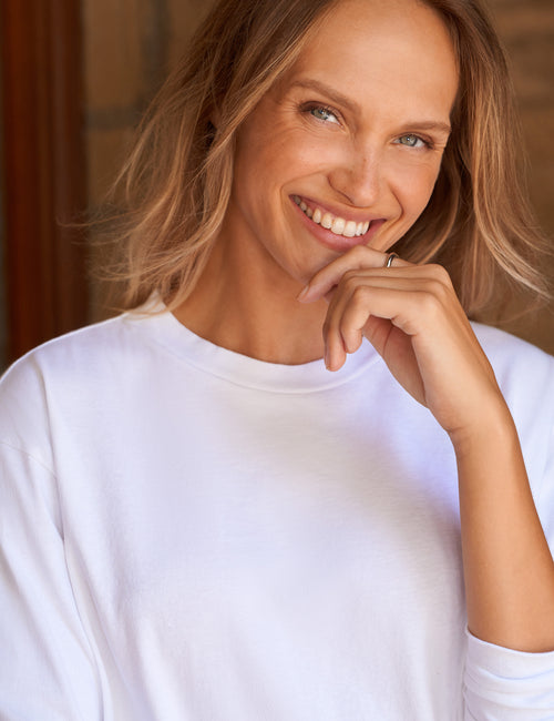 A smiling blonde woman wearing a plain white long-sleeve crewneck shirt, posing with her hand near her chin against a warm, softly blurred background., view 3