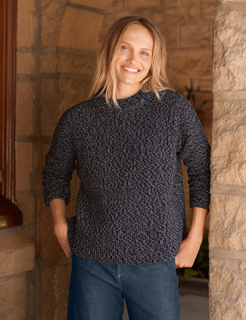 A smiling woman with blonde hair stands in a stone doorway wearing a dark heathered knit sweater with blue jeans., front view