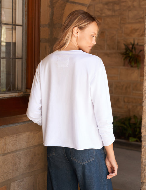 A woman wearing a white long-sleeve top and dark blue jeans, photographed from the back in an outdoor setting with a stone wall and window in the background., alternate view