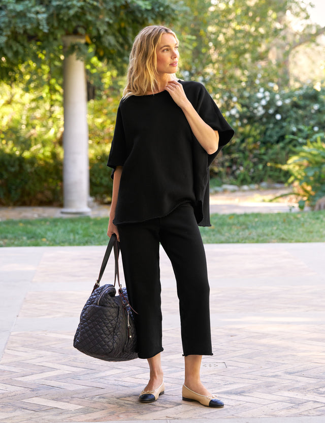 A woman wearing a black olive capelet and black catherine sweatpants standing outdoors with a tote bag and sandals.