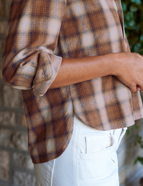 A close-up of a person wearing a brown and beige plaid button-up shirt with rolled-up sleeves, paired with white pants, set against a blurred stone wall background., view 3