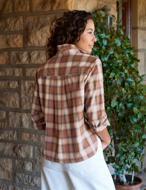 A woman wearing a brown and beige plaid button-up shirt, shown from the back as she stands on a stone porch next to a green potted plant., alternate view