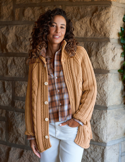 A woman wearing a chunky cable-knit tan cardigan over a brown and white plaid button-down shirt, paired with white pants, standing against a rustic stone wall., front view