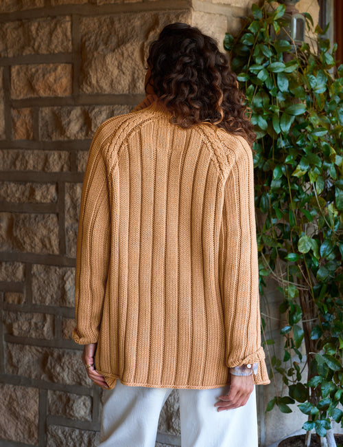 A person viewed from behind wearing a chunky cable-knit sweater in a warm camel tan color, paired with white pants, standing in front of a stone wall and green ivy., alternate view